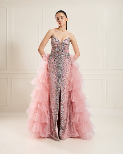 A woman in a strapless, blush-pink gown with crystal embellishments and a voluminous tulle train. The gown features a plunging neckline and a front slit.